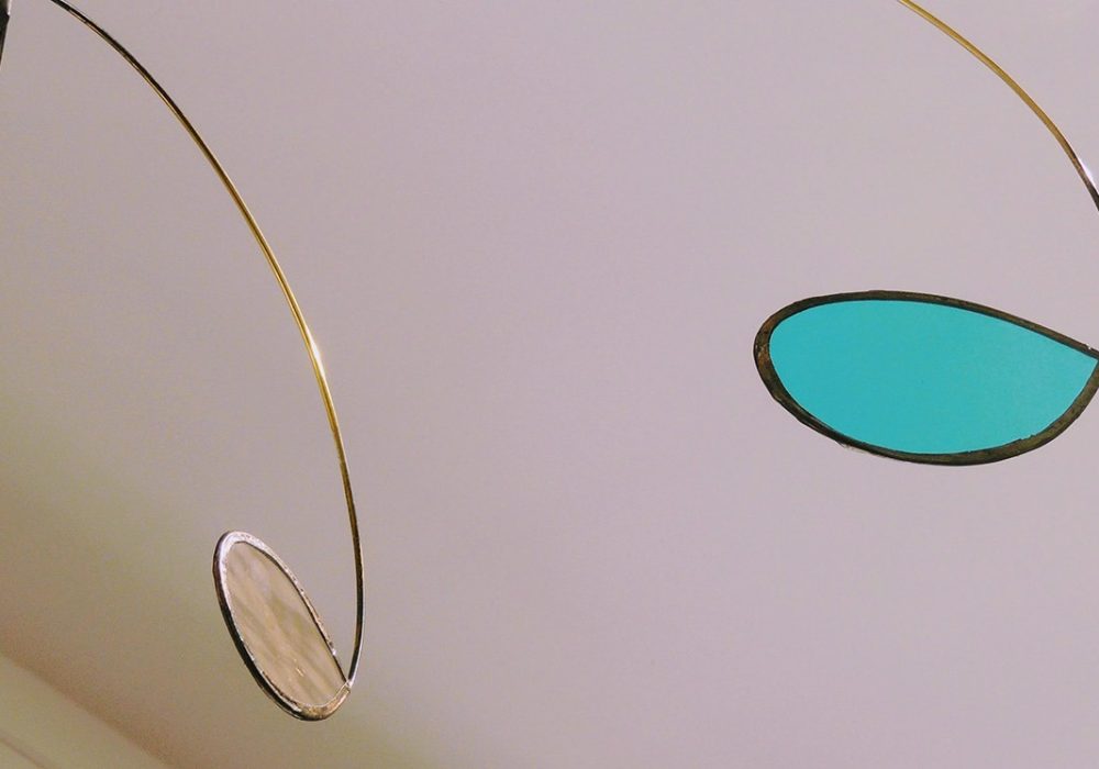 Photographie rapprochée d'un mobile montrant un vitrail bleu ciel découpé en goutte d'eau et serti par une tige de laiton qui prolonge la forme.