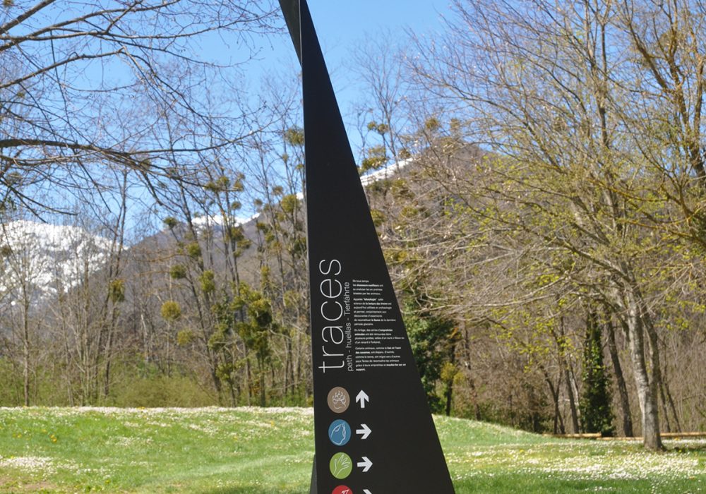 Photographie d'un totem design, en acier plié et thermolaqué, en cohérence avec l'architecture contemporaine marquée du site. Le principe design n'est pas sans rappeler les origamis. Celui-ci, destiné à marquer et informer un lieu d'exposition ou d'animation extérieur, comprend le nom et la description de l'atelier, ainsi que des symboles directionnels.